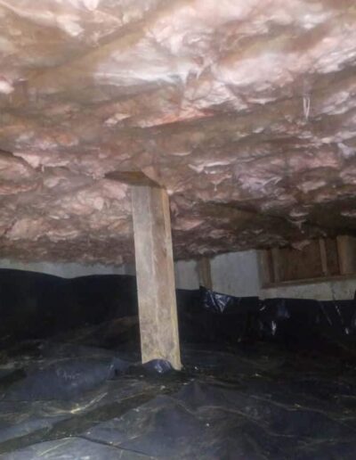 interior of a house's crawl space with insulation installed between the floor joists and a vapor barrier on the ground